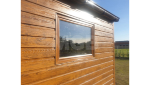 6x3m-es delux fém kerti tároló nyeregtetővel, csokoládébarna cserepeslemez tetővel, világos aranytölgy falakkal, billenőkapuval és ajtóval, ablakkal, ereszcsatornával, szegélyekkel, horganyzott vázszerkezettel, vízszintes lemezborítással